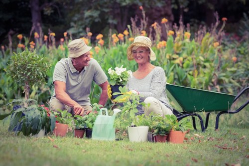 A scene in a garden with two elderly individuals engaging in gardening activities. The man is kneeling on the grass, wearing a beige hat and light grey t-shirt, while the woman, also in a beige hat and light-colored top, is seated on the grass, smiling and holding a potted plant. Around them are various potted plants and gardening tools, including a watering can and a small shovel, on a well-maintained lawn with lush greenery. In the background, there is a vibrant flower border with purple and orange flowers and dense trees, indicating a bright, partly sunny outdoor environment typical of a well-tended UK garden in Bounds Green. A green wheelbarrow is positioned nearby, suggesting ongoing gardening work, aligned with professional garden maintenance and planting services offered by Gardening Services Bounds Green. The scene captures a peaceful, natural setting focused on outdoor gardening activities, plant care, and garden layout.