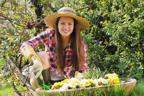 A young woman with long brown hair, dressed in a red and white checkered shirt, wearing gardening gloves and a wide-brimmed straw hat, is kneeling in a lush garden. She is smiling and tending to a flower bed that contains a variety of blooming flowers with white, yellow, and purple petals. Behind her, there is dense green foliage, including trees and bushes, creating a vibrant, natural outdoor environment. The garden features a well-maintained lawn with healthy, dense grass in the foreground, and a wooden garden bed filled with flowers in the middle ground. The scene appears to be during a sunny day, with bright natural light illuminating the garden, highlighting the varied textures of the grass, leaves, and flowers. This setting reflects a tidy and flourishing outdoor space typical of residential gardens in the bounds of London, UK, supporting the services offered by Gardening Services Bounds Green, including flower bed maintenance and general garden care.