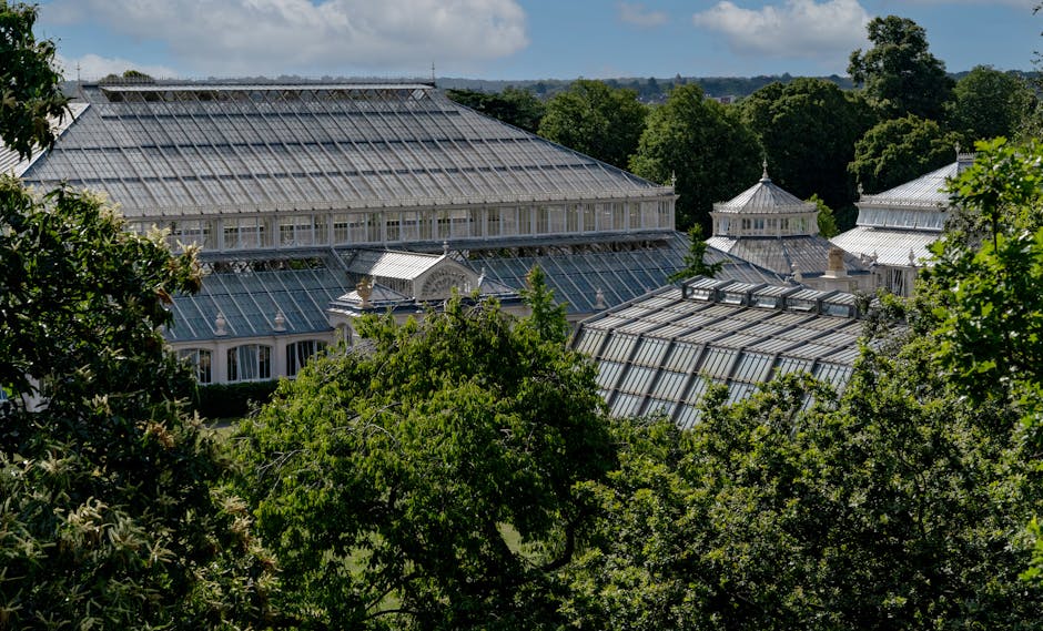 A large Victorian-style glasshouse complex with ornate metal framework and multiple sections, surrounded by lush green trees and garden beds. The glass structures feature peaked roofs and decorative d