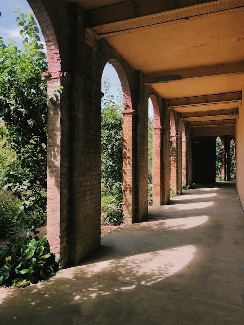 A large Victorian-style glasshouse complex with ornate metal framework and multiple sections, surrounded by lush green trees and garden beds. The glass structures feature peaked roofs and decorative d